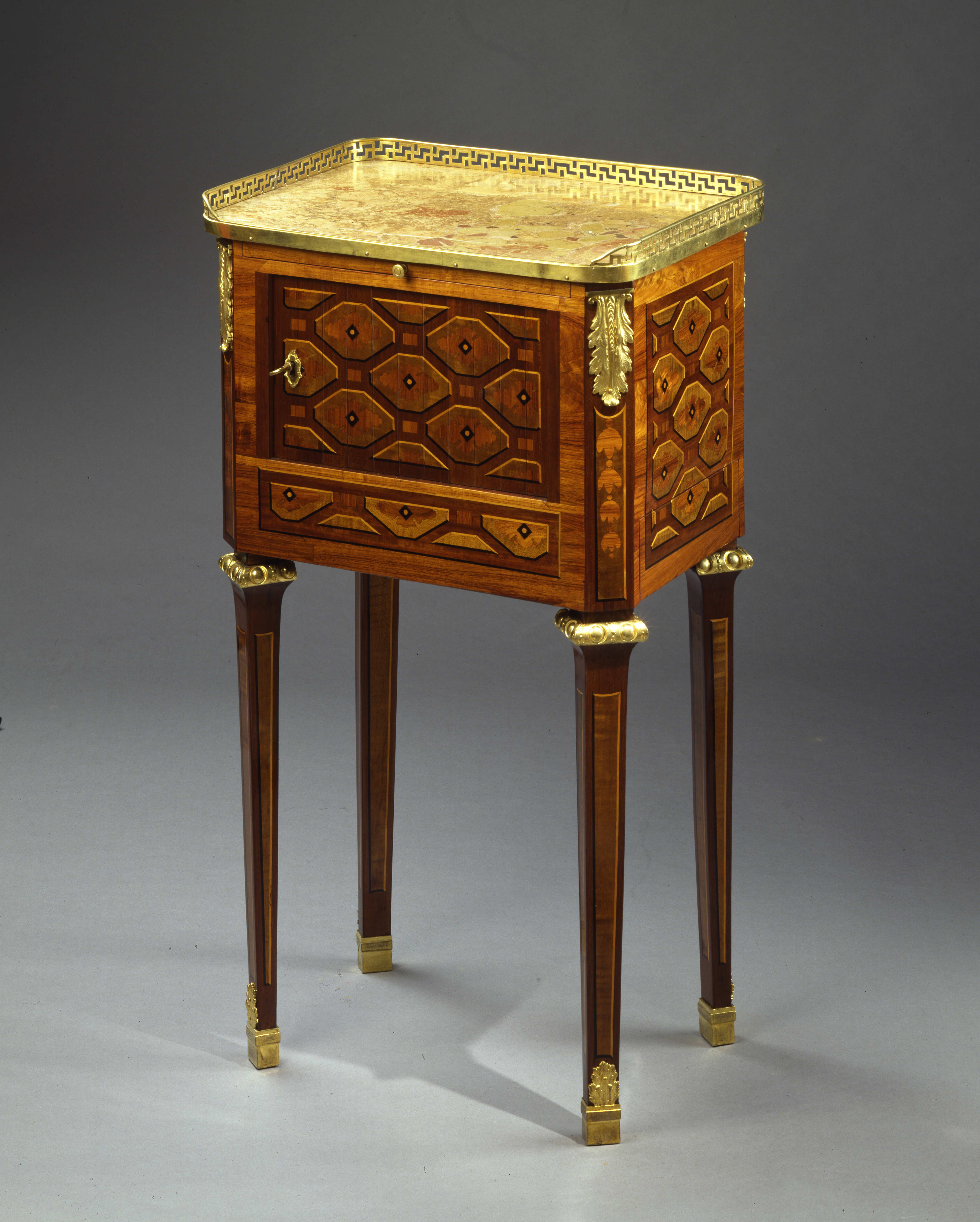 Table marquetée sur toutes ses faces de carrelages octogonaux, ouvrant à une porte à rideau coulissant et un tiroir sur le côté.  Pieds fuselés en gaine.  Ornements de bronze ciselé et doré, galerie, chutes, chapiteaux et sabots. Dessus de marbre de Sienne.