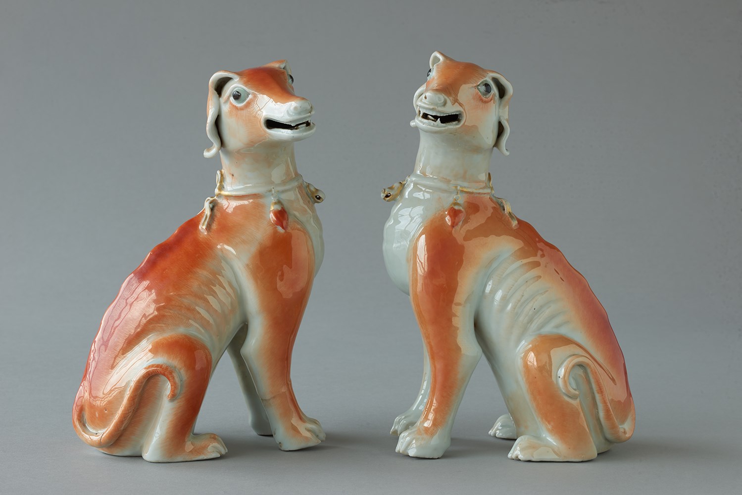 Paire de chiens assis en porcelaine de Chine de commande, émaillés corail au nature. Ils portent autour du cou un collier à grelots dorés.